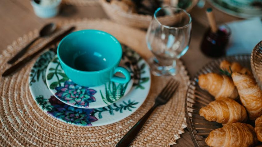 A cozy breakfast setup featuring a teal teacup, plate with floral design, and freshly baked croissants on a woven placemat.